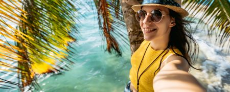 Young beautiful attractive Caucasian woman leaning on a palm trunk by the sea and and making selfies.